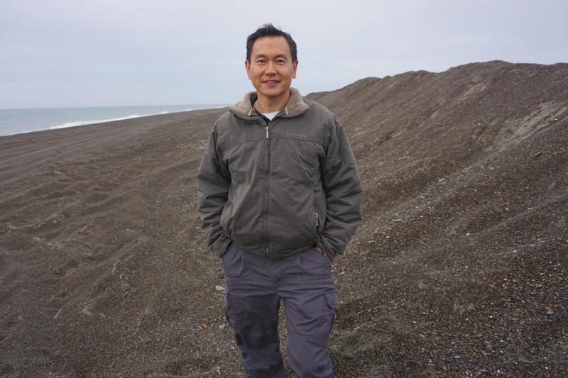 Ming Xiao, an engineering professor at Penn State University, stands on Aug. 3 by a piled-up sand berm used as a makeshift barrier to protect parts of Utqiagvik from storm-driven flooding. Xiao is leading a project that uses a fiber-optic cable to track the minute movements of warming and thawing permafrost. (Photo by Yereth Rosen/Alaska Beacon)